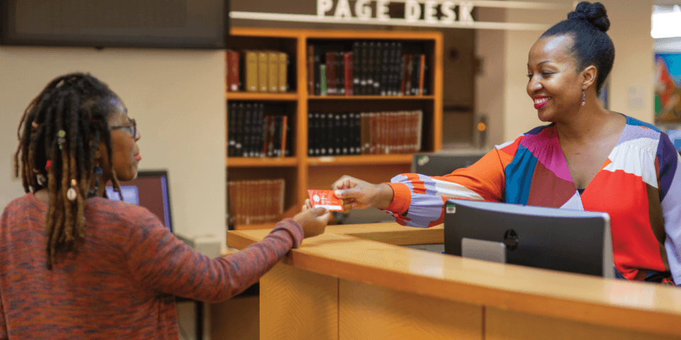 A patron receives a library card from an NYPL staff member at a desk. 