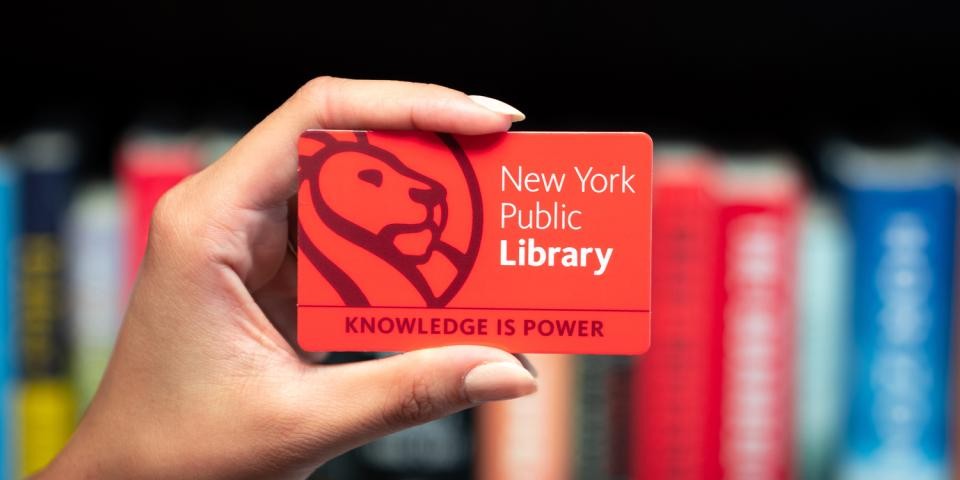 a hand holds a New York Public Library card in front of a bookcase