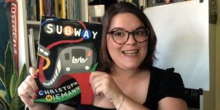 A librarian smiles and holds up a book called Subway by Christoph Niemann