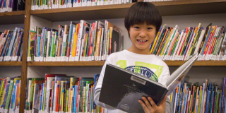 Photograph of a child smiling at the camera while reading a book in front of a library bookshelf filled with books