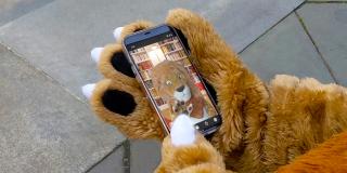 Photo of a lion mascot holding a phone and looking at a screen with the same mascot on it.