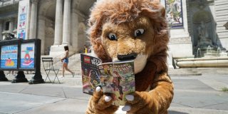 Patience the Lion mascot sits outside of the Stephen A. Schwarzman Building while reading an issue of a Spider-Man comic.