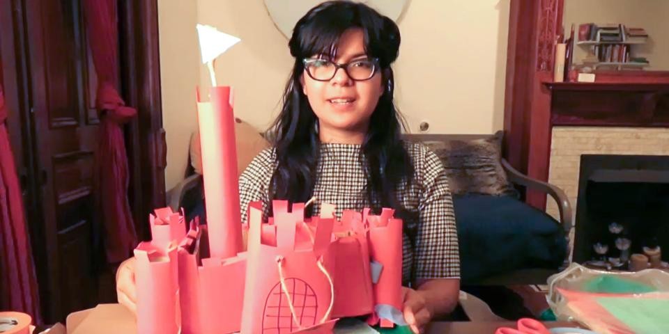 A person with glasses holds up a construction paper castle in a cozy room.