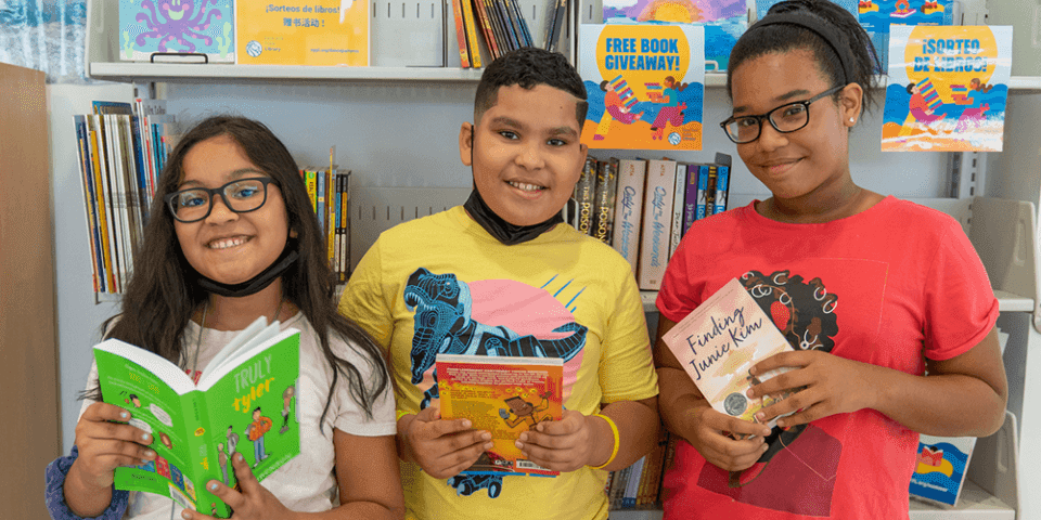 Three kids holding books, standing in front of a wall lined with bookshelves.