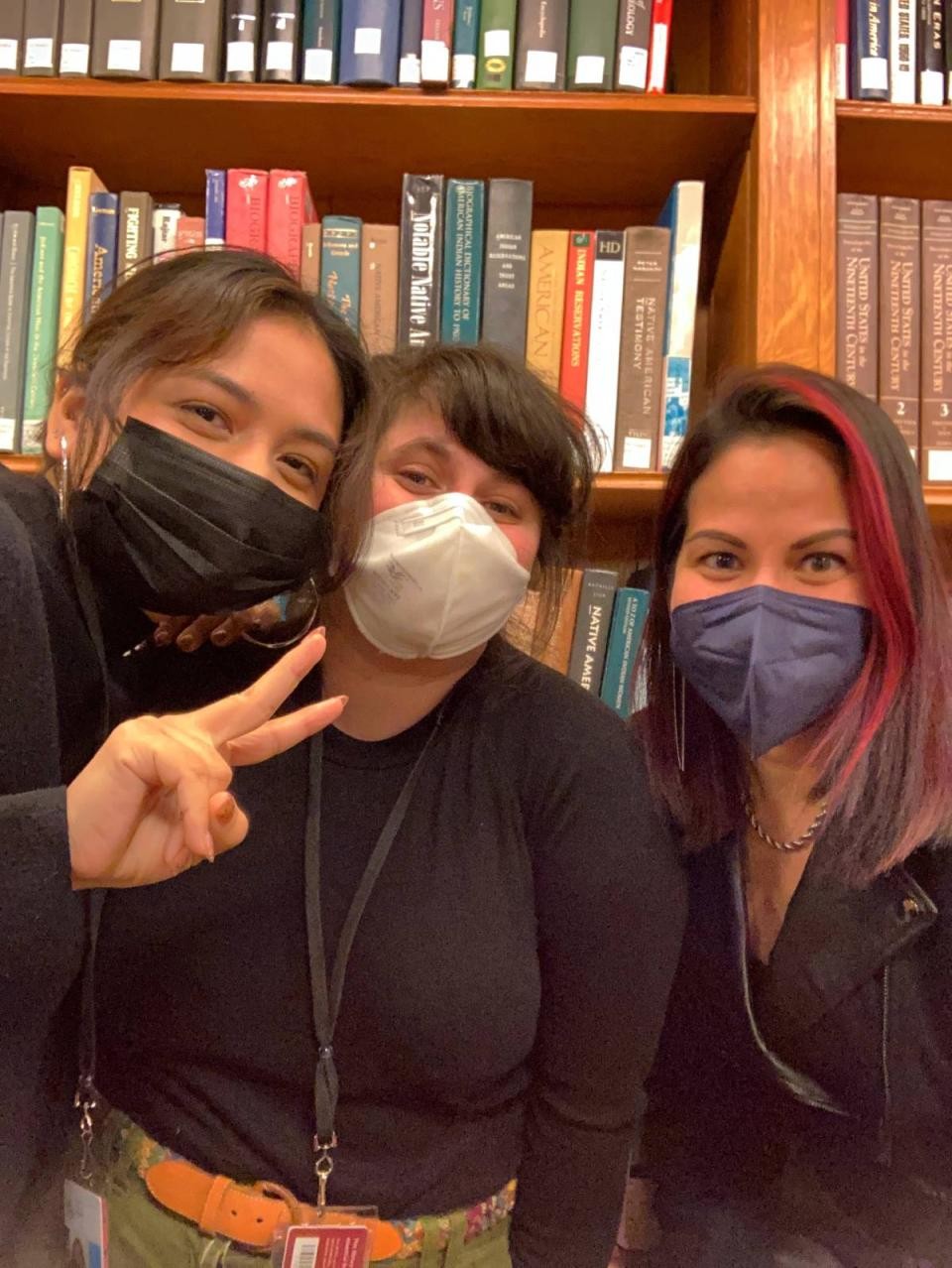 three library staff members, one giving the peace sign, in front of shelves