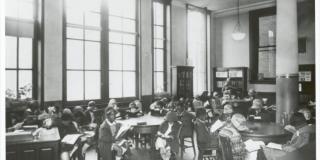 Black and white photograph of a mixed-race group of children in early dress reading in a library.