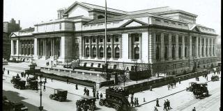 Black and white photograph of the exterior of the Stephen A. Schwarzman Building. Early 20th century cars, a double-decker bus, and a horse-drawn carriage are outside.