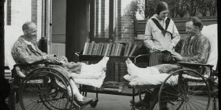 Black and white photograph of two men in wheelchairs each smiling and holding a book. A woman with a library cart stands beside them.