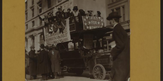 Black-and-white photograph of a bus crowded with women and decked with banners, including one that reads "Votes For Women" and one advertising a rally at Carnegie Hall.