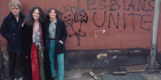 Three women standing in front of a wall that someone has spraypainted "LESBIANS UNITE"