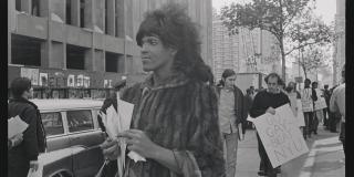 Marsha P. Johnson standing on a New York City sidewalk, wearing a fur coat