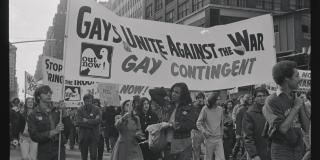 Black and white photo of group marching in a parage, holding a sign saying "Gays Unite Against the War: Gay Contingent"