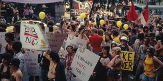 Historic color photograph of a Pride demonstration, featuring people marching with yellow balloons and signs that read: Lesbians Vote Too! 