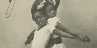 Historic black-and-white photograph of two black children standing back-to-back, smiling, and posing in matching outfits