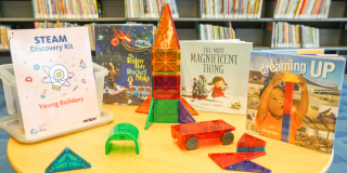 Photo of an NYPL Young Builders Kit spread out on a table with items including: Magna-Tiles Deluxe building set and three books