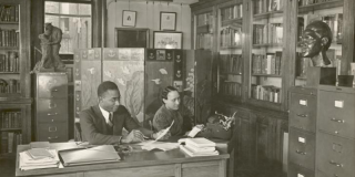 Historic black-and-white photo of Dr. Lawrence D. Reddick and another person sitting at a paper-filled desk in an office full of books, filing cabinets, and sculptures