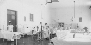 Historic black-and-white photo of a row of beds in a hospital with nurses and doctors milling about nearby