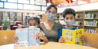 A woman in a face mask and glasses sits at a table, flanked by a young girl and boy who are both masked and holding books. 
