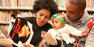 A woman and a man hold a baby girl with a green ribbon and white sweater while reading a picture book titled Cock-a-Doodle-Who!