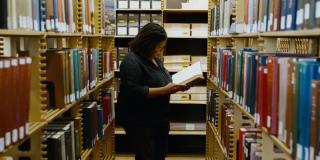 Annette Gordon-Reed, a Black woman with shoulder-length hair wearing black, stands between two rows of bookshelves holding an open book