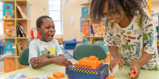 Two kids at a table playing with orange and blue building blocks.