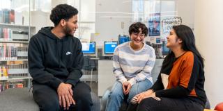 Three teens talking inside a library with computers and bookshelves in the background.
